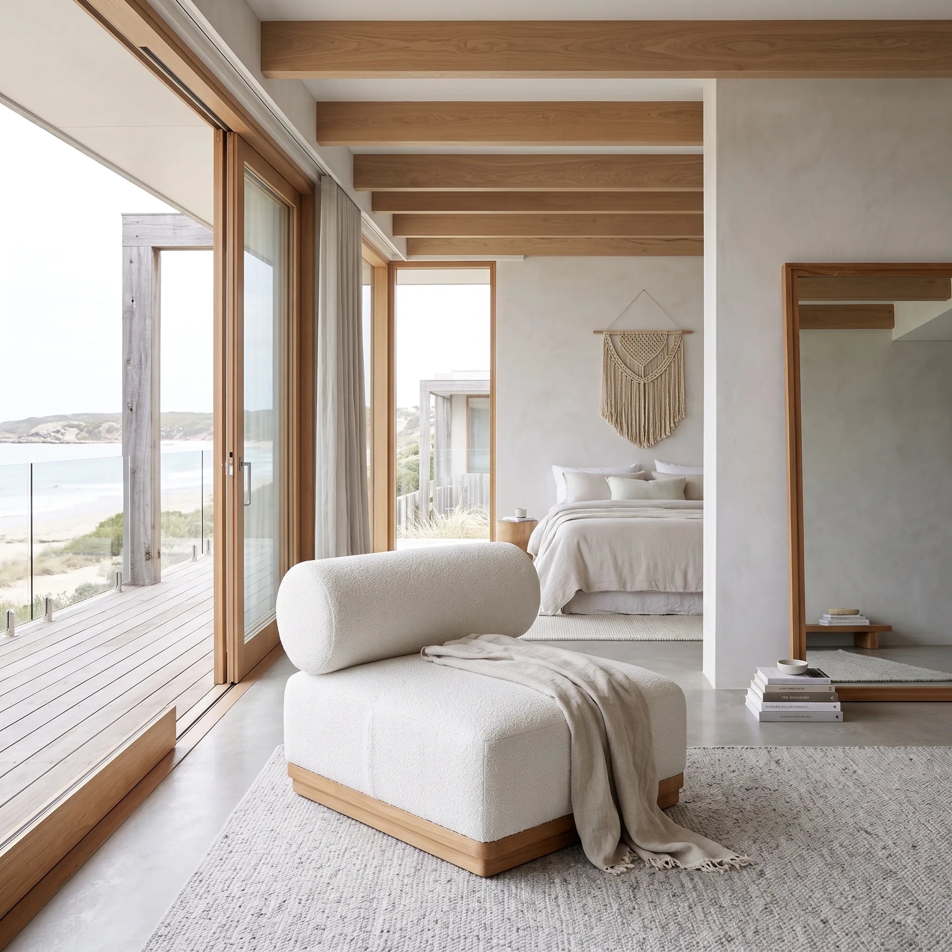 A modern upholstered lounge chair in off-white fabric with a light wood base, styled in a bright, contemporary beachfront bedroom with large windows, wooden beams, and a view of the ocean.