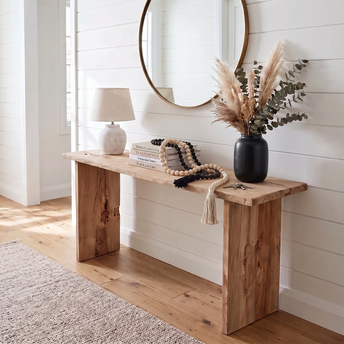A rustic wooden console table styled in a bright, modern entryway with white shiplap walls, a round mirror, a table lamp, books, a beaded rope, and a black vase with dried grasses.