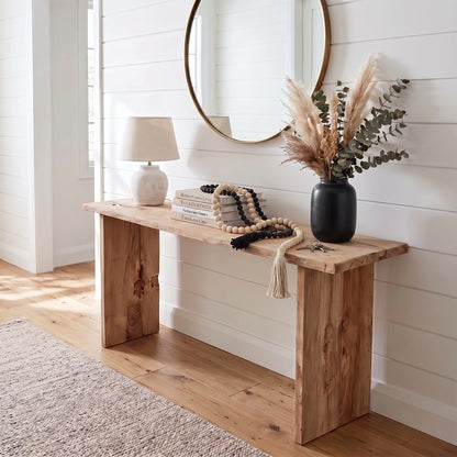 A rustic wooden console table styled in a bright, modern entryway with white shiplap walls, a round mirror, a table lamp, books, a beaded rope, and a black vase with dried grasses.