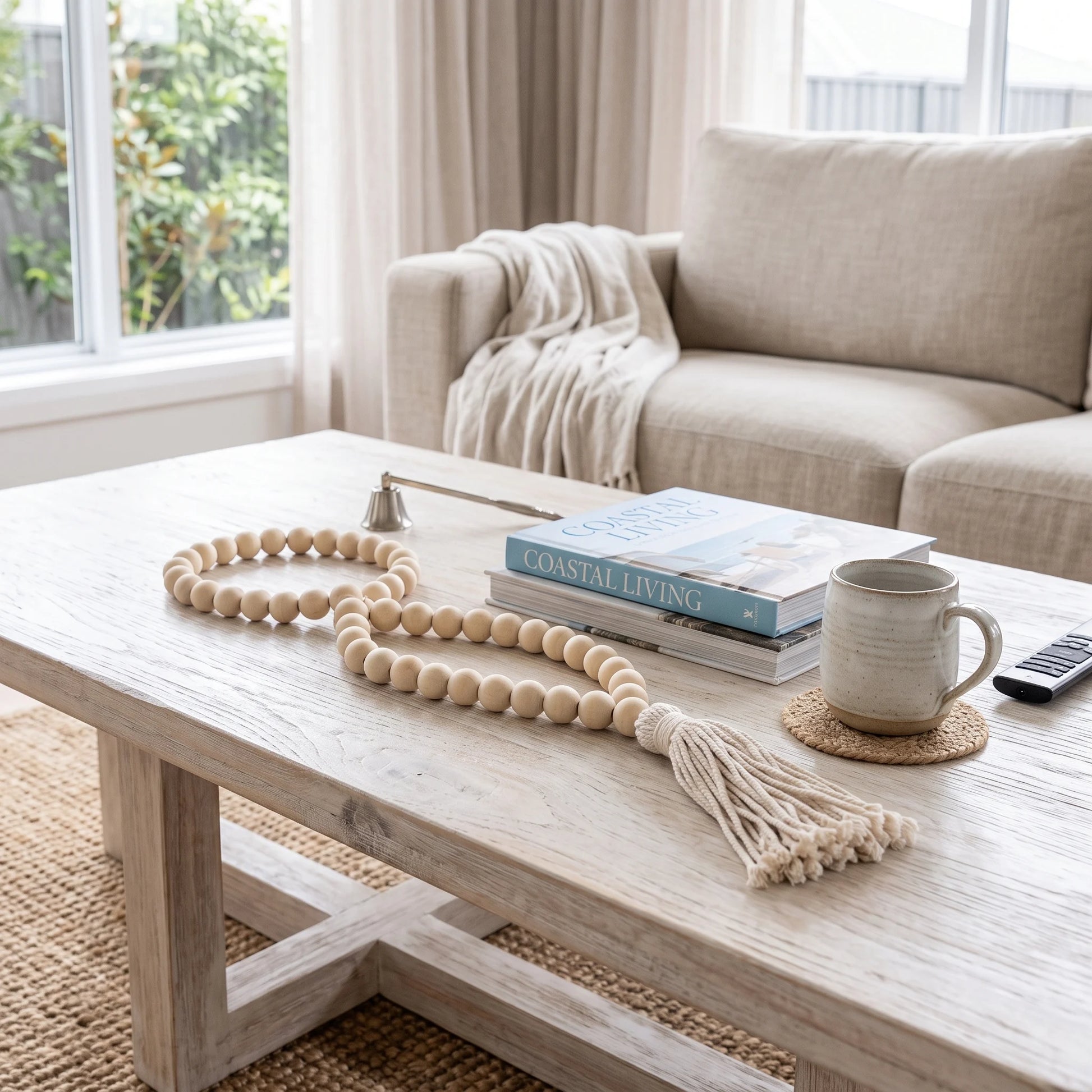 A long strand of light natural wood beads with a tassel lies on a light wood coffee table in a bright living room. The table also holds stacked books, a ceramic mug on a woven coaster, and a small bell.