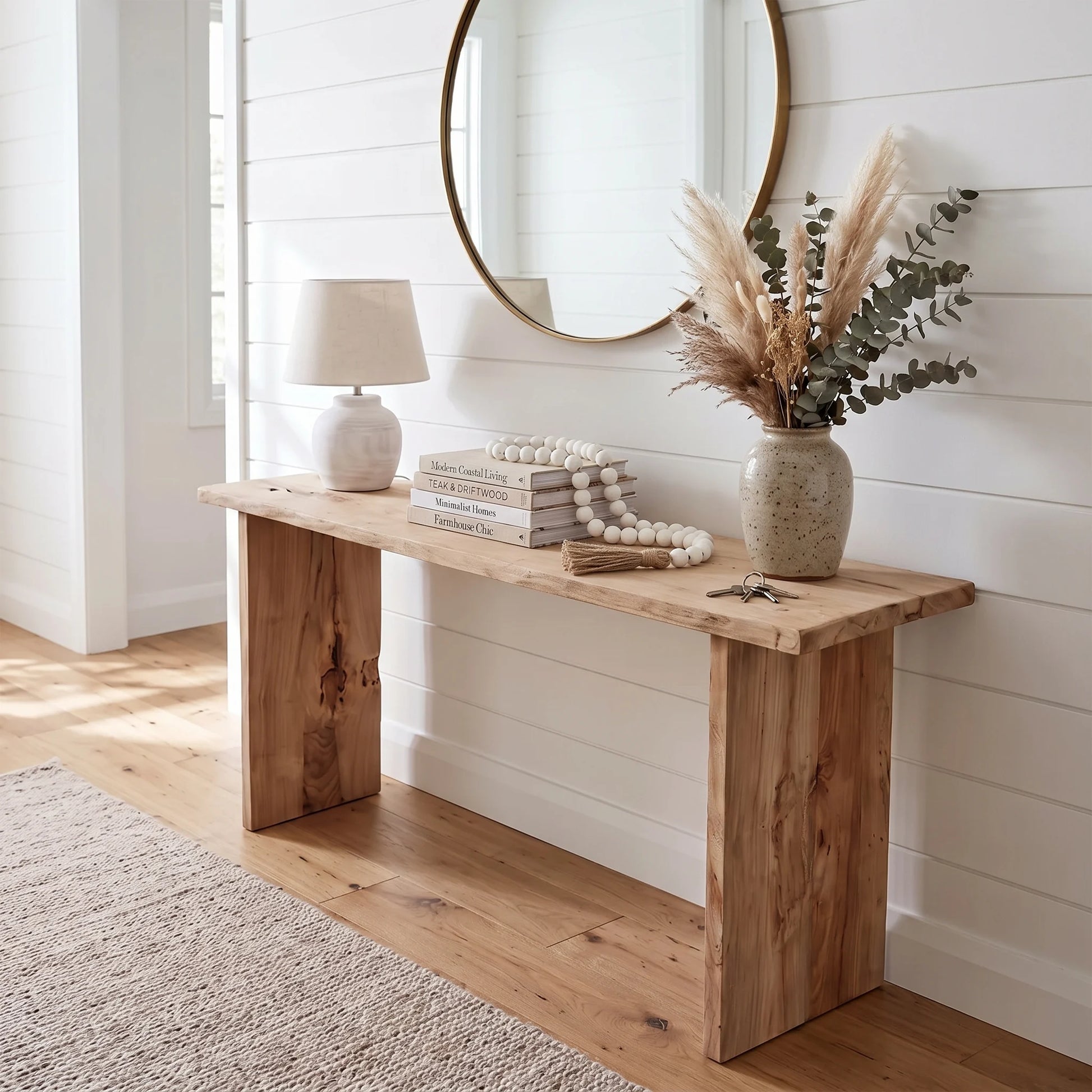 A rustic light brown wooden console table styled in a bright, modern farmhouse entryway with a white shiplap wall, a round mirror, a table lamp, books, and a vase of dried grasses.