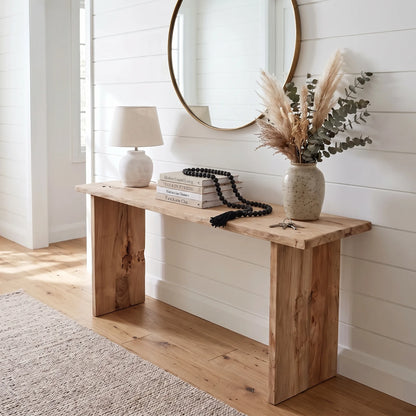 A rustic light brown wooden console table styled in a bright, modern home entryway with white shiplap walls, a round mirror, a white table lamp, books, and a vase of dried pampas grass.