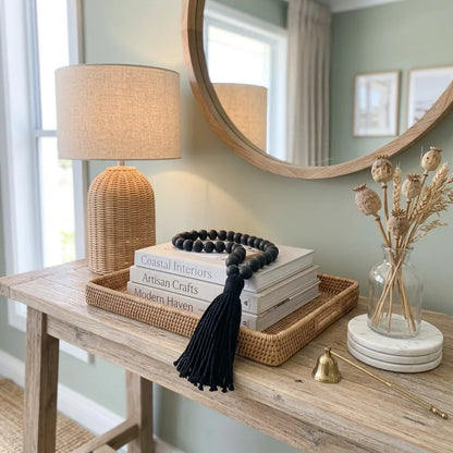 A styled home decor entryway scene featuring a round wooden-framed mirror, a woven rattan table lamp, a stack of books, a black beaded tassel necklace, a wicker tray, and a glass vase with dried grasses on a rustic wooden console table.