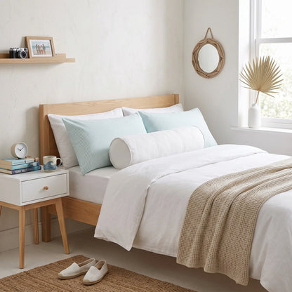 A neatly made bed in a bright, minimalist bedroom. The bed has a light natural wood frame, white bedding, two pale blue pillows, a white cylindrical bolster pillow, and a beige textured throw blanket.