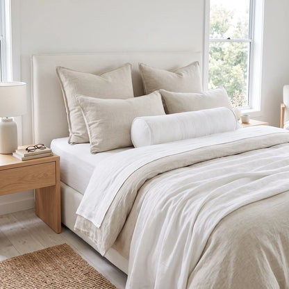 A neatly made bed in a bright bedroom, styled with multiple off-white and beige pillows, a white cylindrical bolster pillow, white bedding, and a light beige duvet. A light wood bedside table with a white lamp and books sits to the left, and a window with greenery outside is visible in the background.