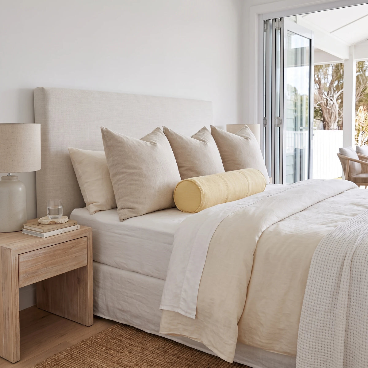 A neatly made bed in a bright, modern bedroom featuring a light beige upholstered headboard, cream bedding, multiple beige pillows, and a pale yellow cylindrical bolster pillow. A light wood nightstand with a matching lamp sits beside the bed, and sliding glass doors open to a balcony.