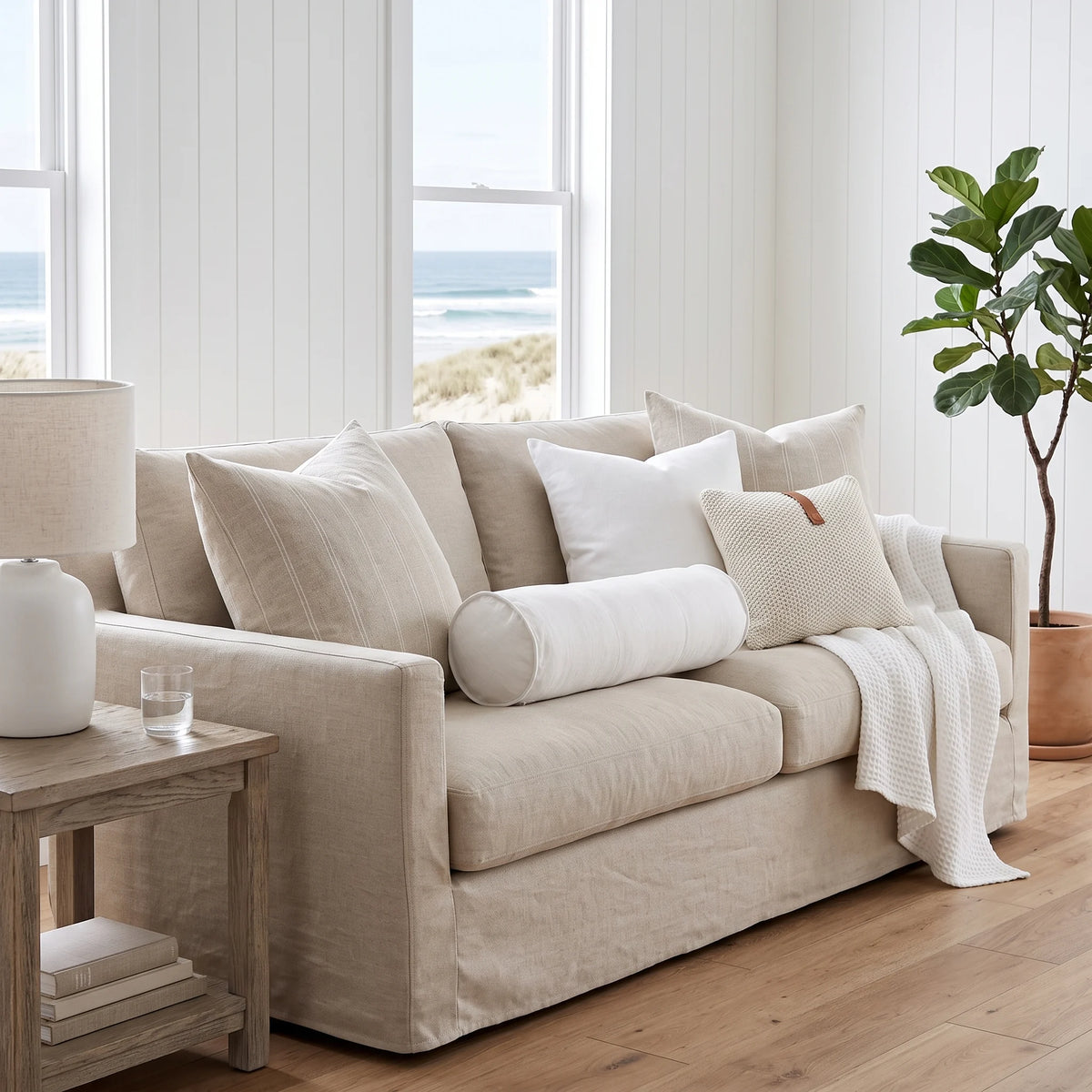 A beige upholstered loveseat in a bright coastal living room, styled with white and beige pillows, a white throw blanket, a wooden side table with a lamp, and a potted plant near large windows overlooking the beach.