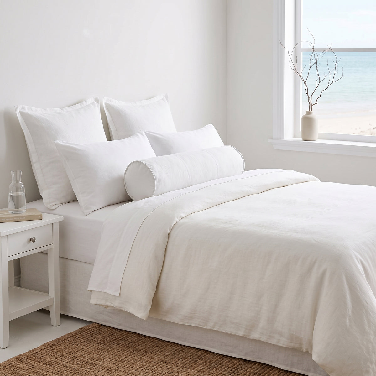 A neatly made bed with white linens and multiple pillows in a bright, minimal bedroom. A white nightstand sits beside the bed, and a window looks out to the ocean with a small vase of branches on the sill.
