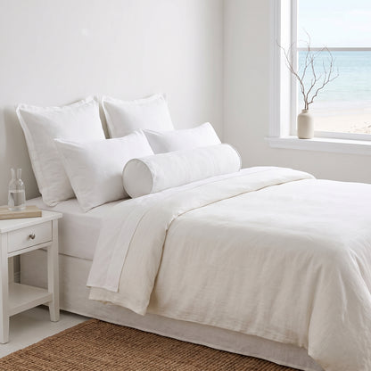 A neatly made bed with white linens and multiple pillows in a bright, minimal bedroom. A white nightstand sits beside the bed, and a window looks out to the ocean with a small vase of branches on the sill.