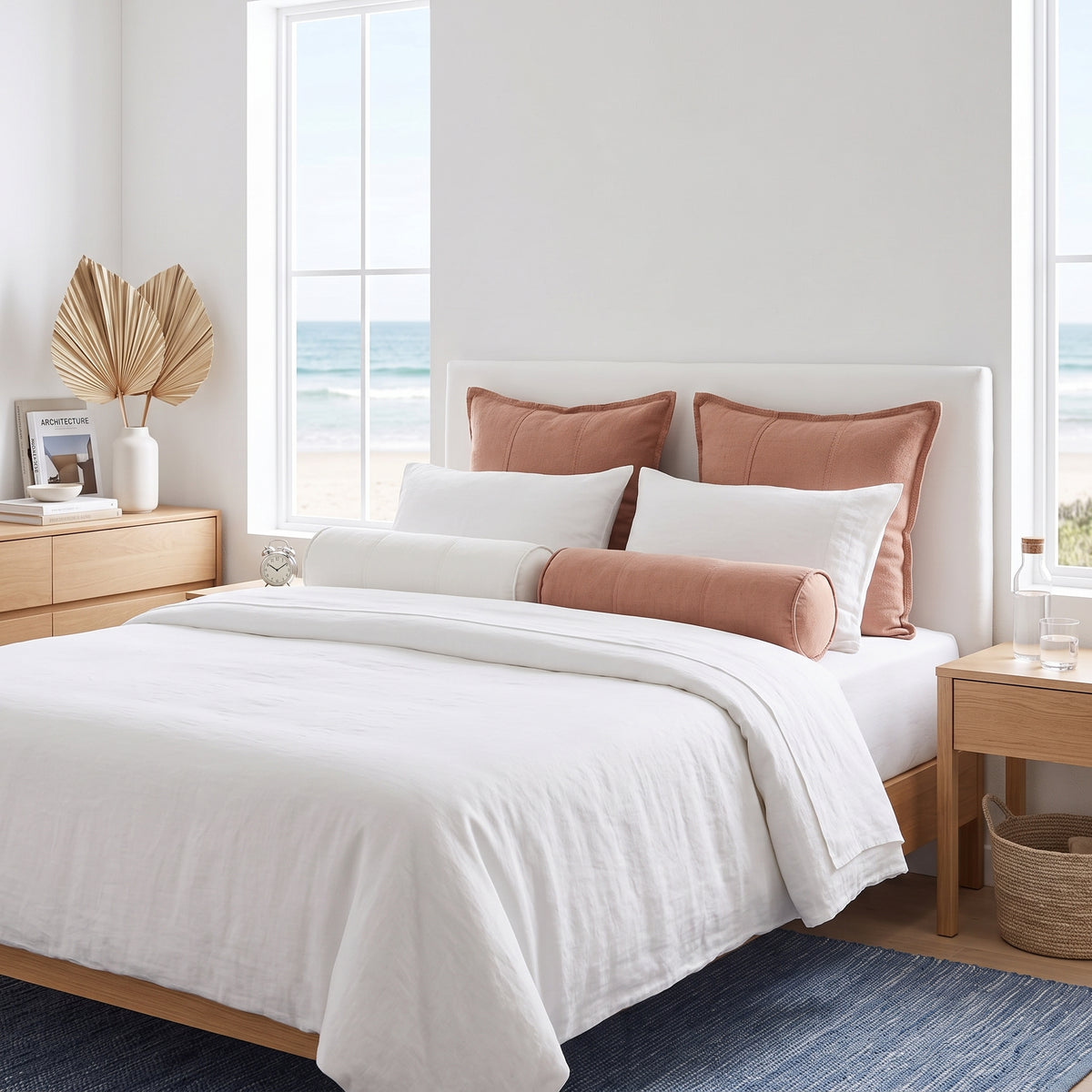 A neatly made bed with white bedding and blush pink pillows, styled in a bright beach-view bedroom with wooden nightstands and large windows.