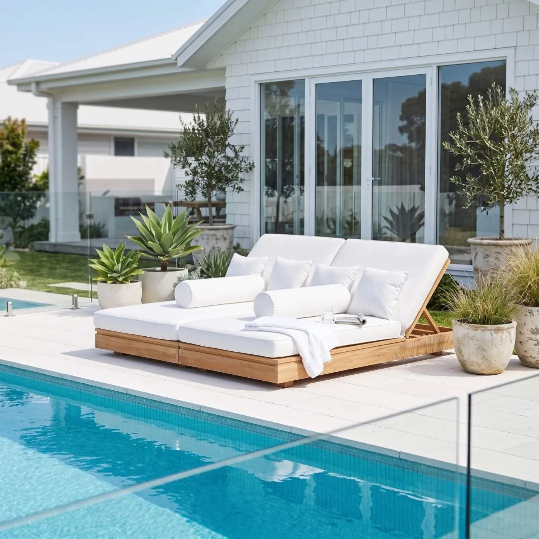 A modern outdoor daybed with a light wood frame and thick white cushions sits beside a swimming pool on a tiled patio, with a white house and potted plants in the background.