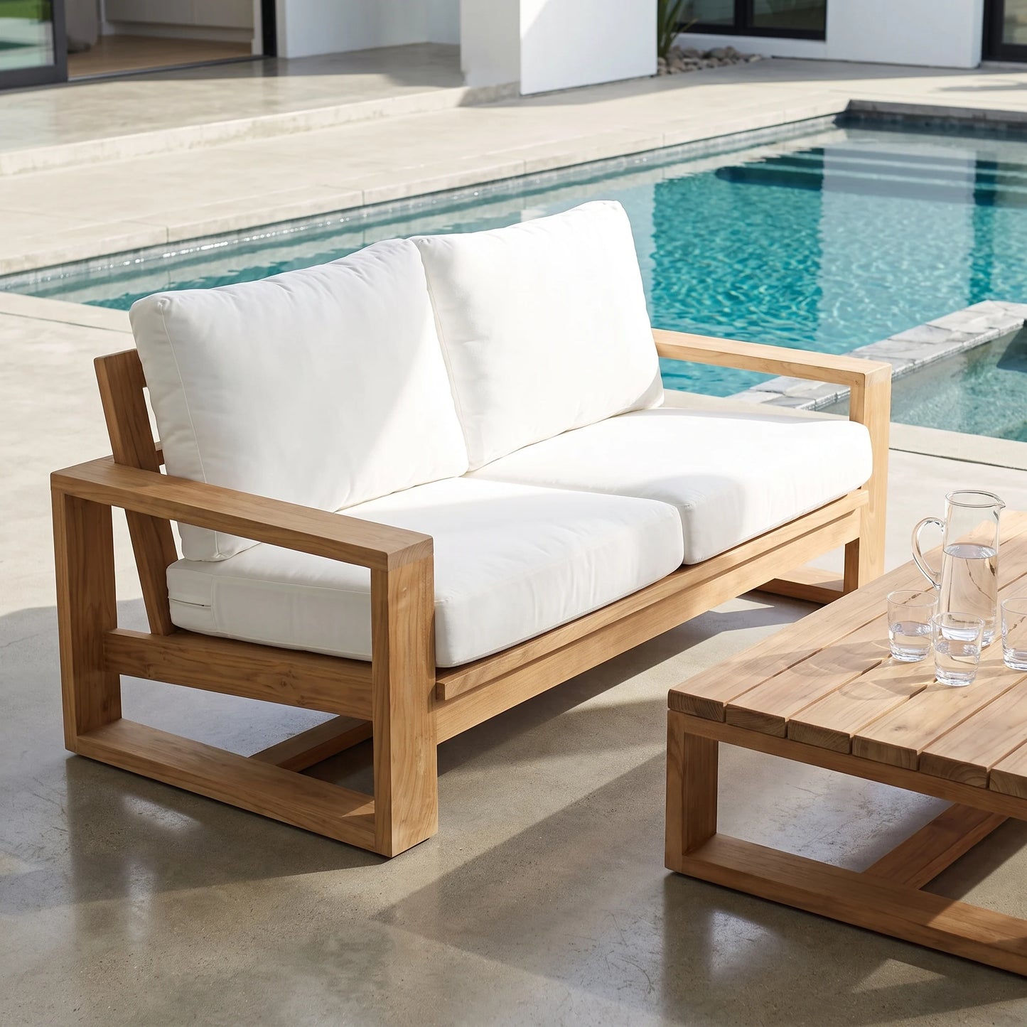 A two-seat outdoor sofa with a natural wood frame and thick white cushions, positioned on a concrete patio next to a swimming pool and a matching wooden coffee table.