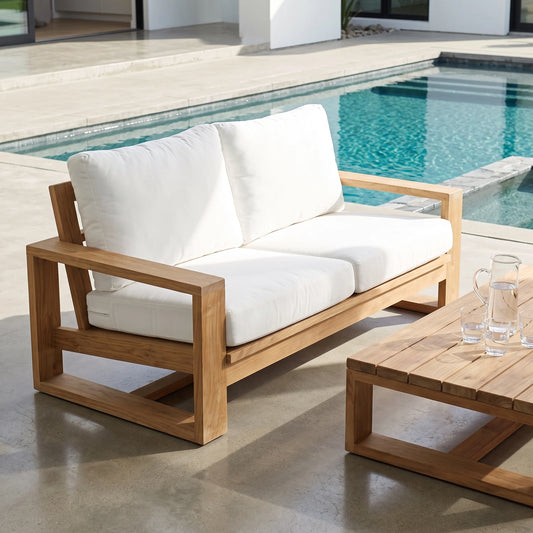 A two-seat outdoor sofa with a natural wood frame and thick white cushions, positioned on a concrete patio next to a swimming pool and a matching wooden coffee table.