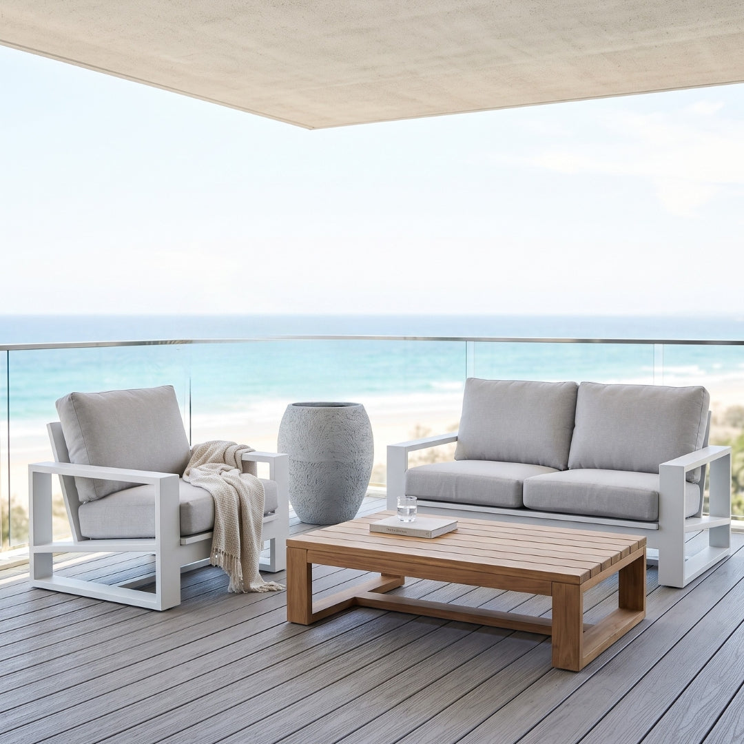 Outdoor patio furniture set on a wooden deck overlooking the ocean, featuring a white-framed armchair with a beige throw, a matching white-framed sofa with light gray cushions, and a rectangular wooden coffee table.