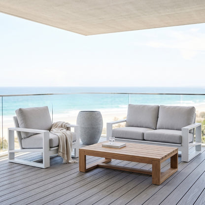 Outdoor patio furniture set on a wooden deck overlooking the ocean, featuring a white-framed armchair with a beige throw, a matching white-framed sofa with light gray cushions, and a rectangular wooden coffee table.