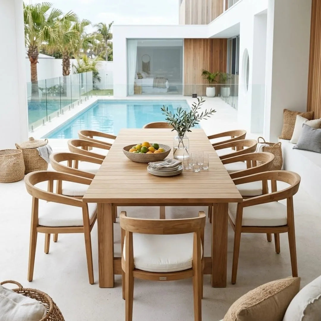 A modern dining set with a rectangular light wood table and matching curved-back chairs with white cushions, styled in a bright, contemporary outdoor patio setting next to a swimming pool.