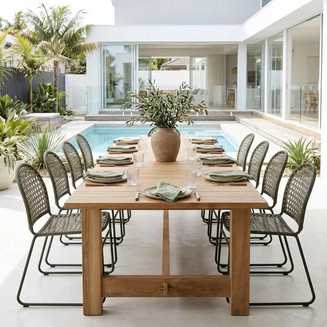 A rectangular wooden dining table set with twelve woven metal chairs, place settings, and a large greenery centerpiece, positioned outdoors on a patio next to a swimming pool and modern house.