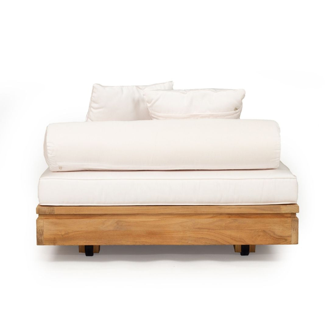 A modern outdoor daybed featuring a natural teak wood frame, a thick cream-colored mattress, a matching rolled lumbar pillow, and two square back pillows, shown against a solid white background.