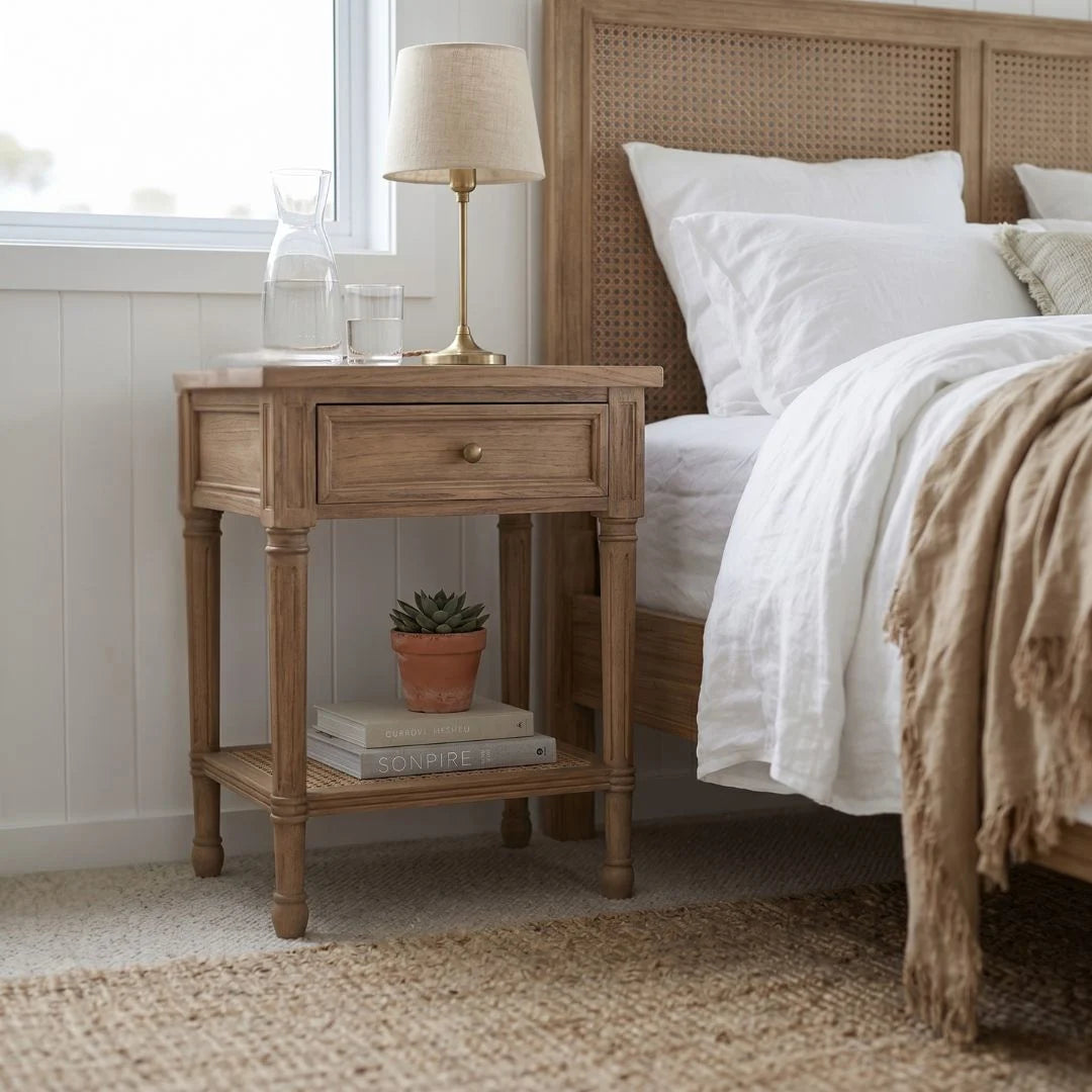 Wooden nightstand with lamp and books next to a bed with white bedding and a woven headboard.