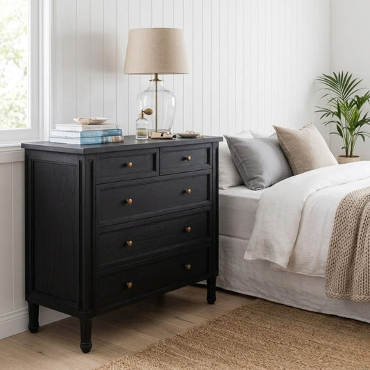 Black dresser with gold knobs next to a bed with gray and beige pillows in a bedroom.