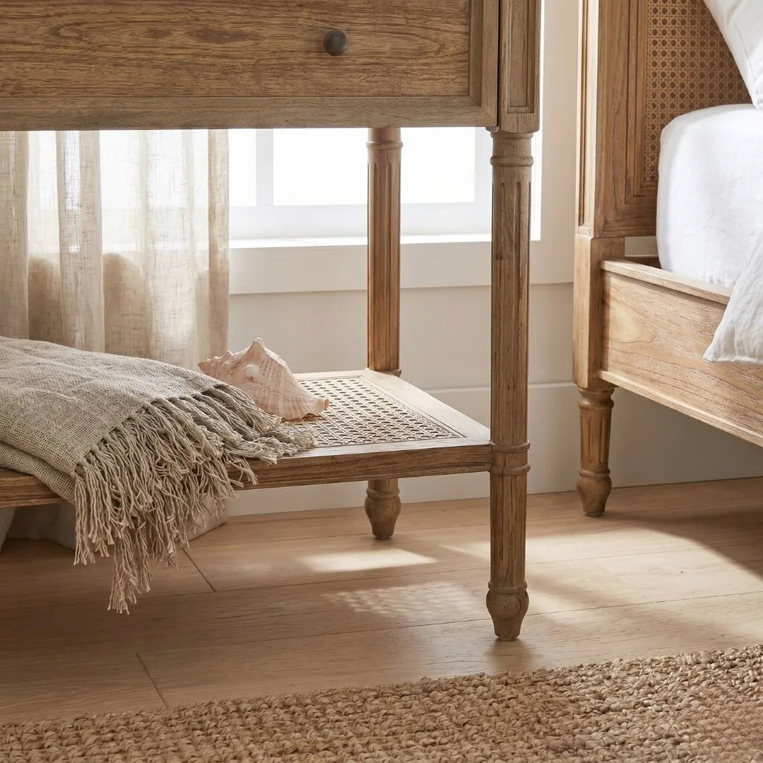 Wooden side table with a textured blanket and pillow next to a bed in a softly lit room.