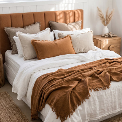 A styled bed with white bedding, multiple beige and rust accent pillows, a rust throw blanket draped across the foot, and a tufted rust headboard in a bright bedroom setting.
