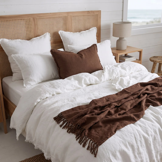 A styled bed in a bright bedroom featuring white linens, a brown throw pillow, and a brown fringed blanket, set against a light wood cane headboard with a bedside lamp and window view.
