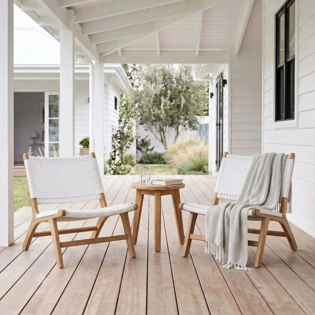 Two light wood outdoor lounge chairs with white woven seats and backs, arranged on a wooden porch with a small round wooden side table between them. A white throw blanket is draped over one chair, and a glass and book sit on the table.