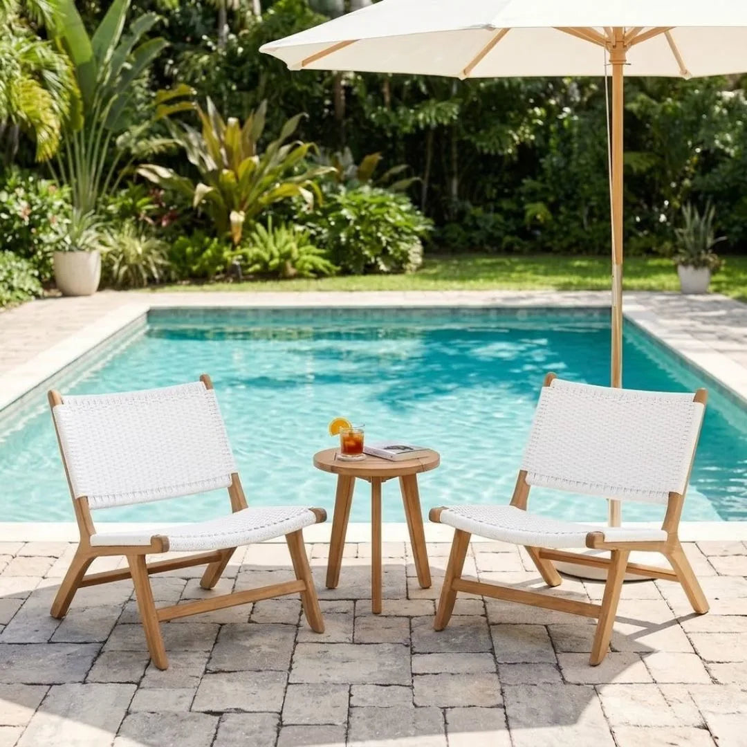 Two outdoor lounge chairs with white woven seats and light wood frames sit on a stone patio beside a swimming pool, with a small round side table between them and a large cream patio umbrella overhead.