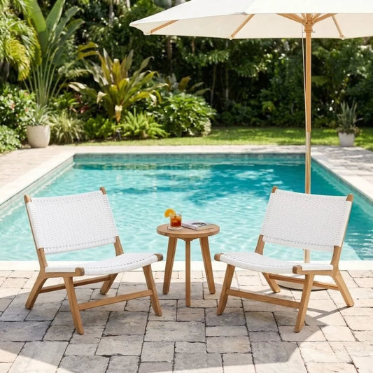 Two outdoor lounge chairs with white woven seats and light wood frames sit on a stone patio beside a swimming pool, with a small round side table between them and a large cream patio umbrella overhead.