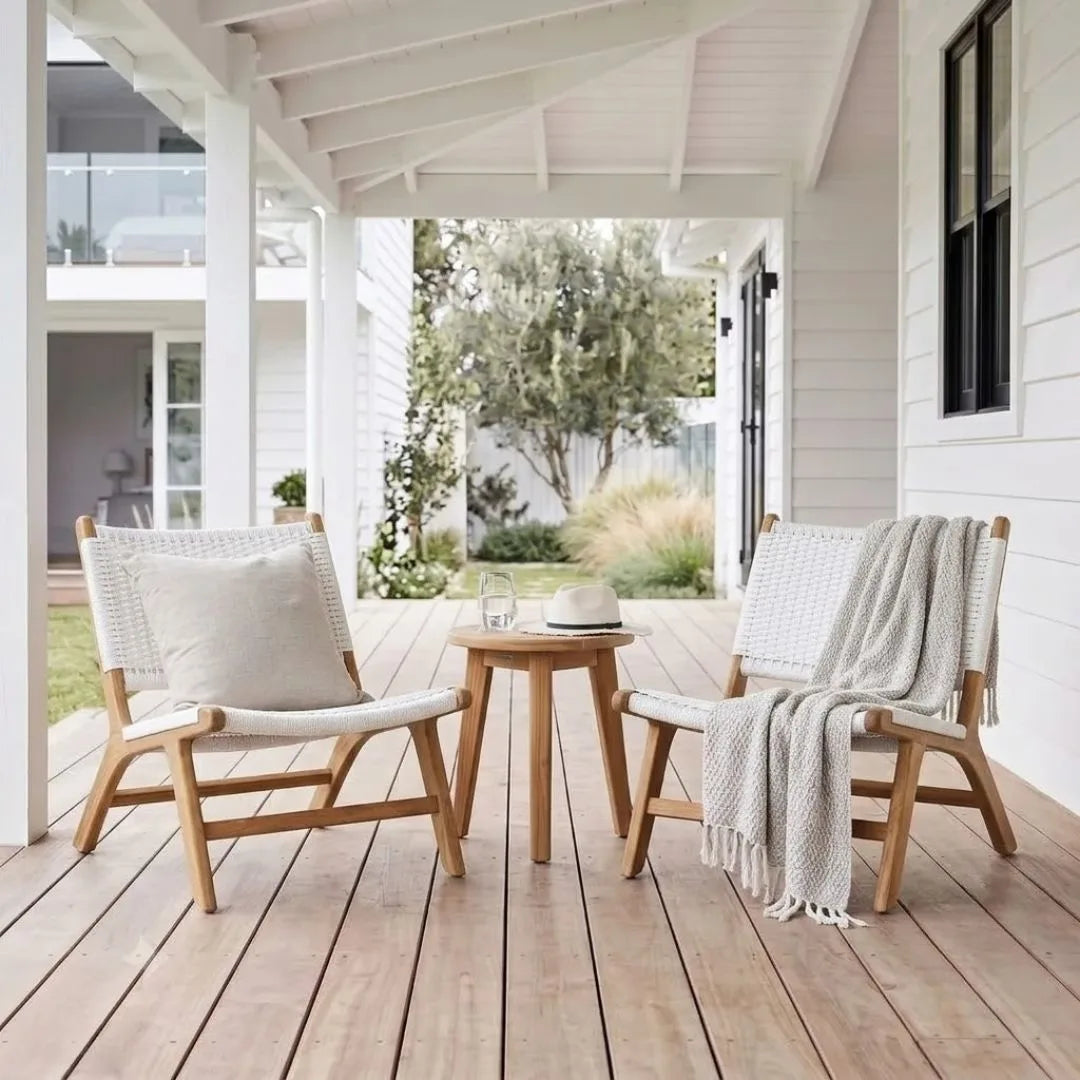 Two light wood outdoor lounge chairs with woven cream cushions are arranged on a wooden porch with a small round wooden side table between them. A white throw blanket is draped over the right chair, and the scene is photographed from above in a bright, airy home setting.