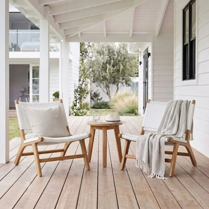 Two light wood outdoor lounge chairs with woven cream cushions are arranged on a wooden porch with a small round wooden side table between them. A white throw blanket is draped over the right chair, and the scene is photographed from above in a bright, airy home setting.