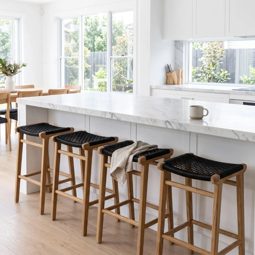 Four wooden bar stools with black woven seats lined up along a white marble kitchen island in a bright, modern kitchen.