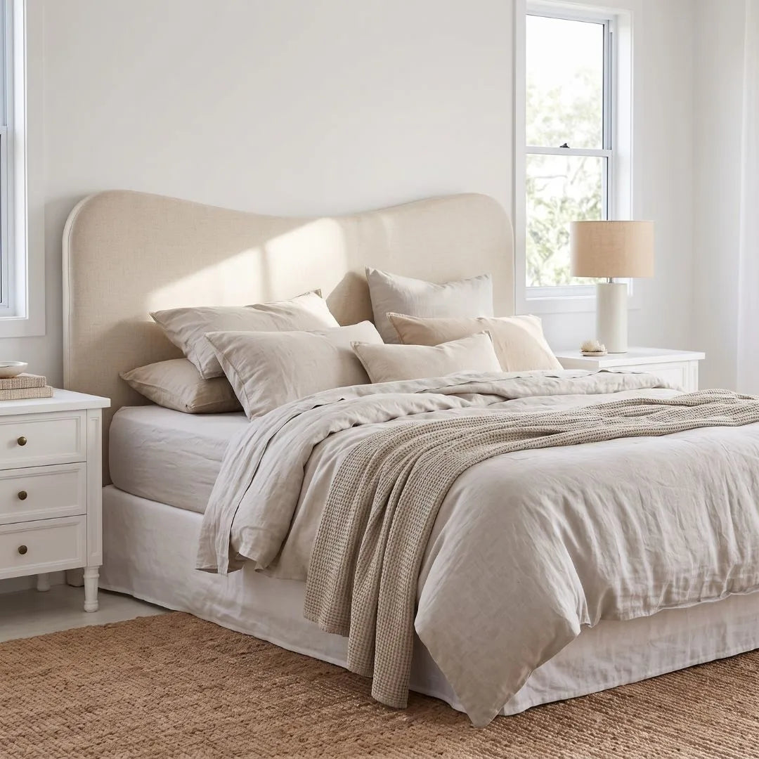 A neatly made bed in a bright, minimalist bedroom featuring a tall upholstered headboard, neutral beige bedding, matching pillows, a textured throw blanket, white nightstands, and a window.