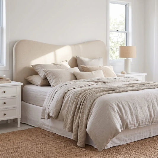 A neatly made bed in a bright, minimalist bedroom featuring a tall upholstered headboard, neutral beige bedding, matching pillows, a textured throw blanket, white nightstands, and a window.