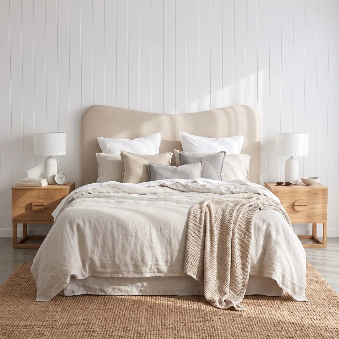 A neatly made bed in a bright bedroom, featuring a beige upholstered headboard, layered neutral bedding in cream, beige, and light gray, flanked by two light wood nightstands with white table lamps.