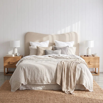 A neatly made bed in a bright bedroom, featuring a beige upholstered headboard, layered neutral bedding in cream, beige, and light gray, flanked by two light wood nightstands with white table lamps.