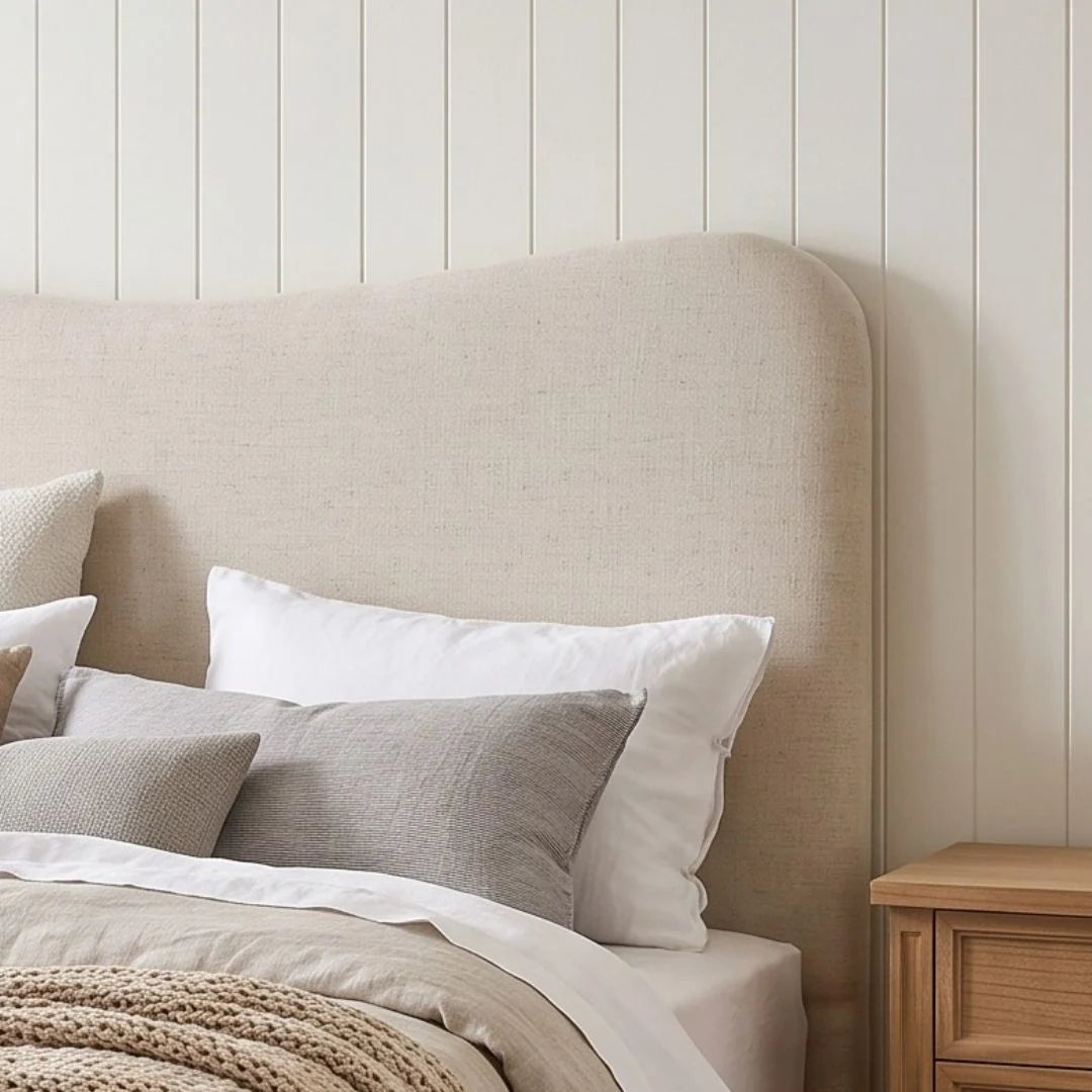 A beige upholstered headboard in a styled bedroom setting, shown with white and gray pillows, neutral bedding, and a light wood nightstand against a white shiplap wall.