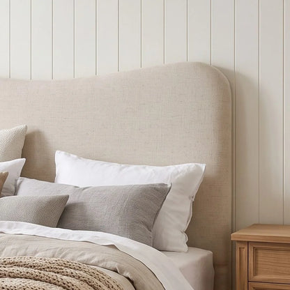 A beige upholstered headboard in a styled bedroom setting, shown with white and gray pillows, neutral bedding, and a light wood nightstand against a white shiplap wall.