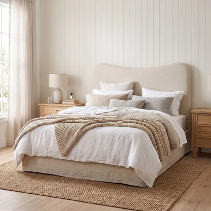 A neatly made bed in a bright, neutral-toned bedroom. The bed has a tall upholstered headboard, white bedding, several light gray pillows, a beige throw blanket, and a woven jute rug on a light wood floor.