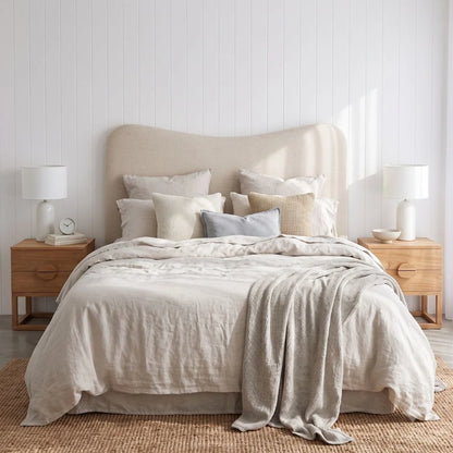 A neatly made bed with light beige linens, multiple neutral and pale blue pillows, a beige upholstered headboard, two wooden nightstands, white table lamps, and a woven rug in a bright bedroom setting.