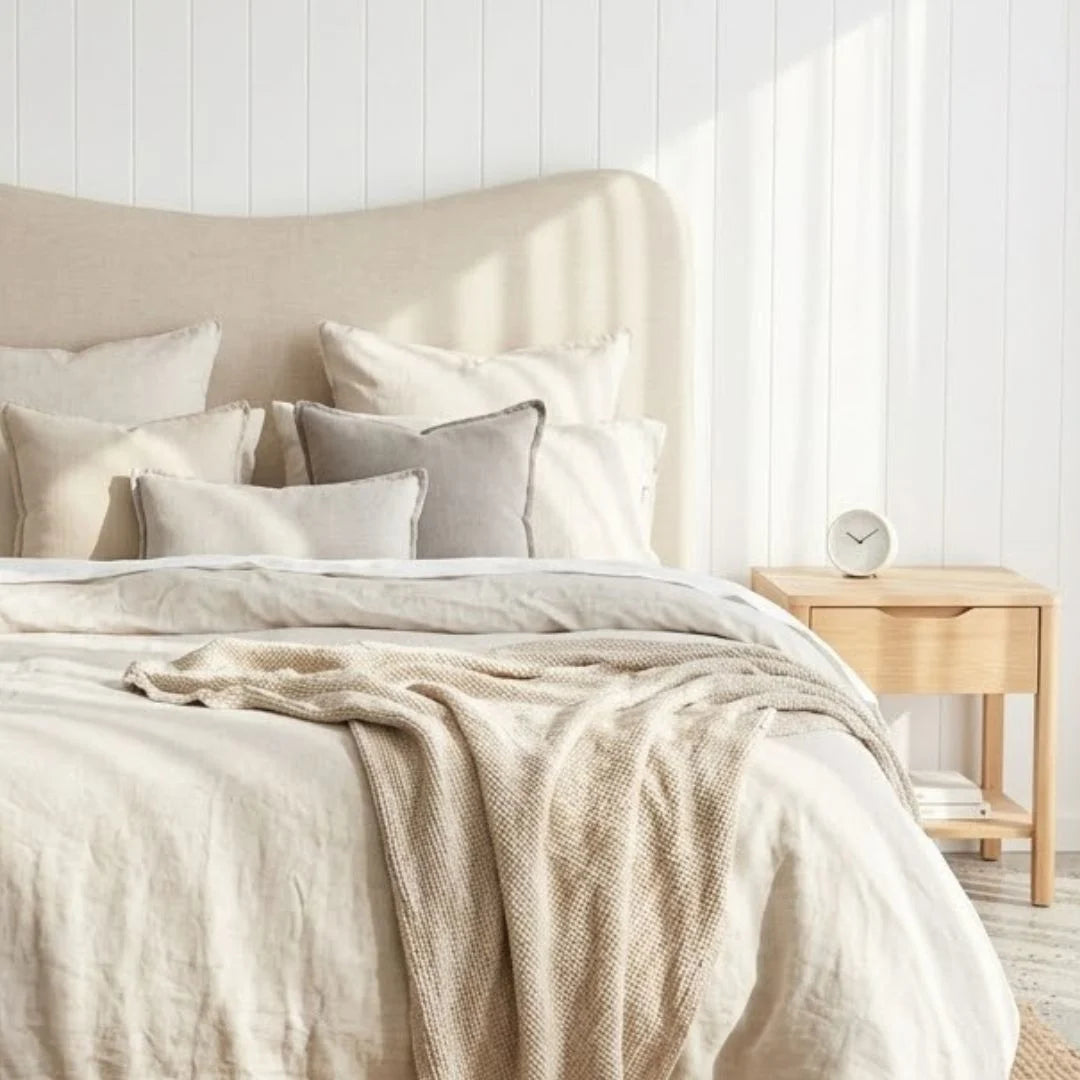 A neatly made bed with cream and beige linens, multiple pillows, a beige upholstered headboard, and a matching textured throw blanket. A light wood nightstand with a small white clock sits beside it against a white paneled wall.