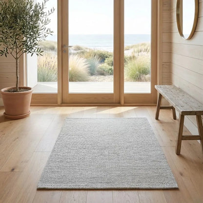 Room with a off white eco rug, wooden bench, and large windows overlooking a garden.