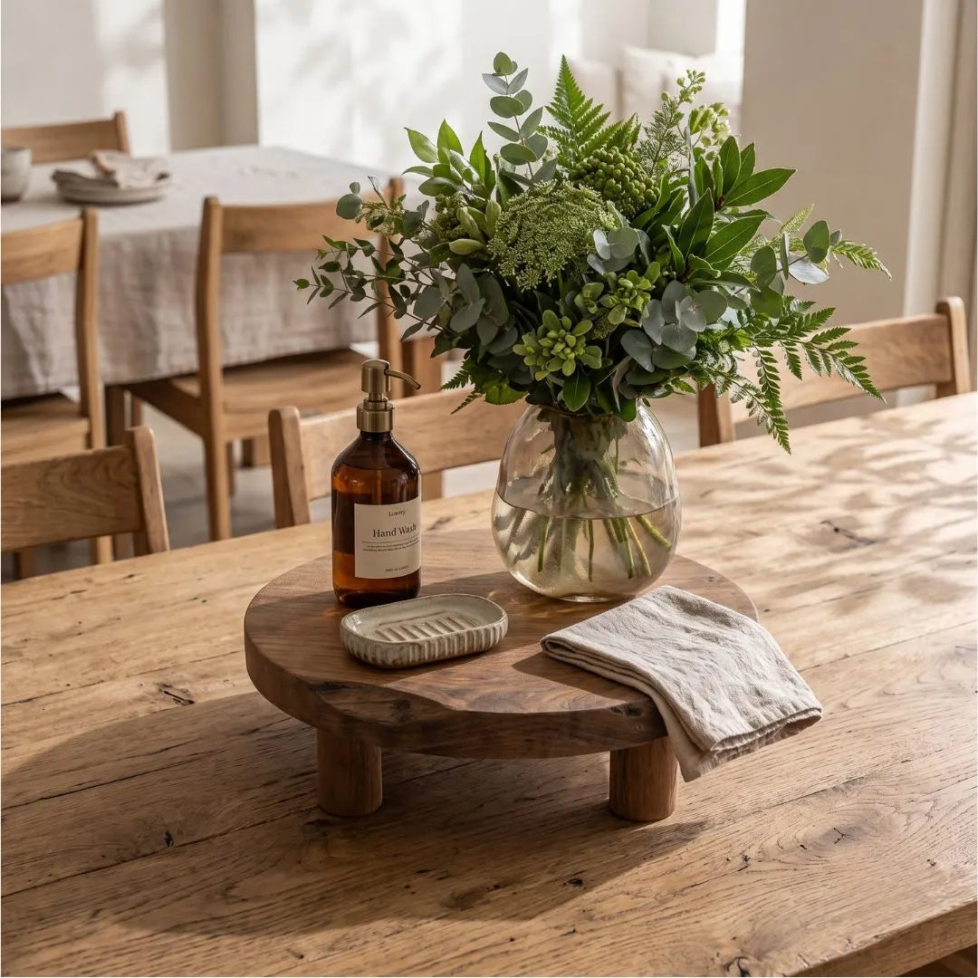 A round wooden tabletop riser holding a clear glass vase with a green floral arrangement, a brown pump bottle, a small scrubber, and a folded linen cloth, set on a rustic wooden dining table in a softly lit room.