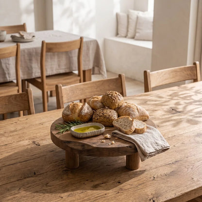 A round wooden bread stand on legs holding several rustic loaves of bread, sliced bread, a small dish of olive oil, and a folded cloth, set on a wooden dining table in a warm, sunlit room.