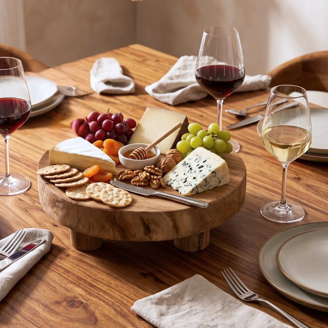 A round wooden charcuterie board styled on a dining table with cheese, crackers, nuts, grapes, and wine glasses around it.