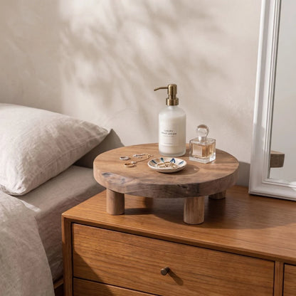 A round wooden nightstand with chunky legs sits beside a bed, styled with a white pump bottle, a small glass perfume bottle, jewelry, and a dish on top. The scene is lit by warm natural light with soft shadows on a beige wall.