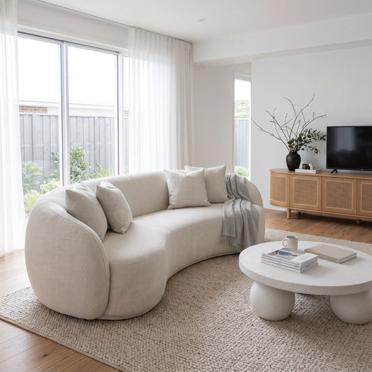 A modern curved upholstered sofa in a bright, minimalist living room with light wood flooring, a round white coffee table, and a large window with sheer white curtains.