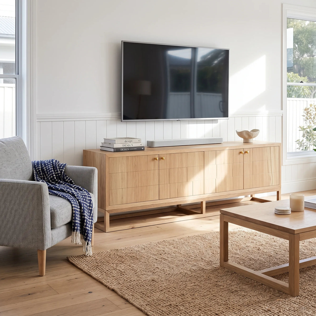 A light natural wood TV stand cabinet in a bright living room, styled with a wall-mounted flat-screen TV, a soundbar, books, and a decorative bowl. The room features a gray armchair with a blue-and-white throw, a woven jute rug, and a wooden coffee table.