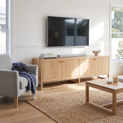 A light natural wood TV stand cabinet in a bright living room, styled with a wall-mounted flat-screen TV, a soundbar, books, and a decorative bowl. The room features a gray armchair with a blue-and-white throw, a woven jute rug, and a wooden coffee table.